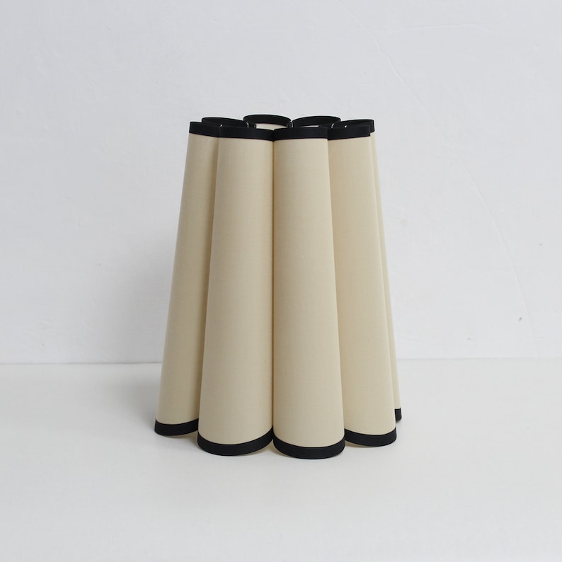 May include: A cream-colored lampshade with a black trim. The lampshade is pleated and has a rounded shape.