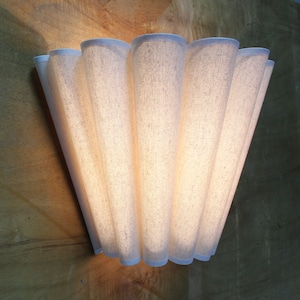 May include: A wall-mounted light fixture with a fan-like design. The fixture is made of a light-colored, textured fabric, with a white trim. The light shines through the fabric, creating a warm glow. The fixture is mounted on a wooden surface.