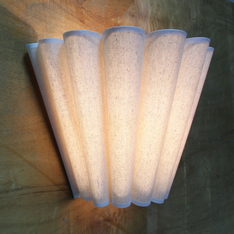 May include: A wall-mounted light fixture with a fan-like design. The fixture is made of a light-colored, textured fabric, with a white trim. The light shines through the fabric, creating a warm glow. The fixture is mounted on a wooden surface.