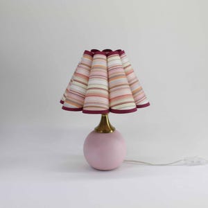 May include: A small, pink table lamp with a striped lampshade. The lampshade features horizontal stripes in shades of pink, orange, and beige, with a dark red trim. The base is a round, light pink shape with a brass-colored neck.