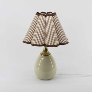 Könnte beinhalten: Eine cremefarbene Tischlampe mit einem braun-weiß karierten Lampenschirm. Der Lampenschirm hat einen gewellten Rand und ist mit braunem Stoff eingefasst. Der Lampenfuß hat eine Tropfenform mit einem goldfarbenen Metallhals.