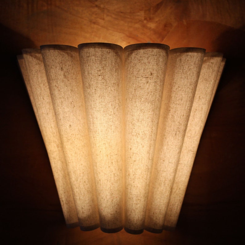 May include: A beige fabric lampshade with a pleated design. The lampshade has a series of vertical, curved panels that create a fan-like shape. The fabric appears to be textured, and the light shines through the material, creating a warm glow.
