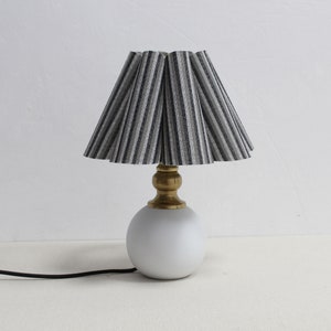 May include: A small table lamp with a white, spherical base and a brass-colored stem. The lampshade is pleated with vertical stripes in shades of gray and black. A black power cord extends from the base.