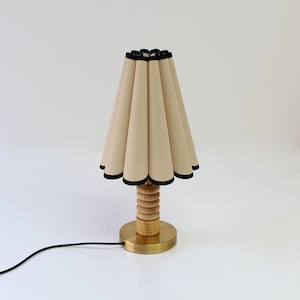 Può includere: Una lampada da tavolo beige con un paralume in tessuto plissettato bordato di nero. La lampada ha una base in legno con un fondo in ottone.
