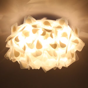 Pode incluir: Uma luminária de teto branca feita de muitas peças translúcidas em forma de lágrima que se sobrepõem. A luz está acesa e ilumina a sala.