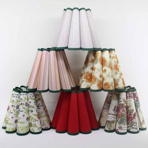 Könnte beinhalten: Eine Sammlung von kegelförmigen Lampenschirmen in verschiedenen Mustern und Farben. Die Schirme umfassen Weiß mit gewebter Textur, rosa und orangefarbene Streifen, florale Designs und einen einfarbigen roten Schirm. Jeder Schirm ist mit grünen Kanten versehen.