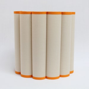 May include: A cylindrical lampshade with a cream-colored fabric exterior and bright orange trim. The shade is composed of vertical, rounded panels, creating a textured surface. The design is modern and minimalist, suitable for various interior styles.