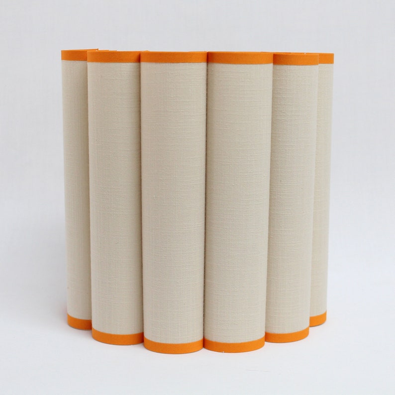 May include: A cylindrical lampshade with a cream-colored fabric exterior and bright orange trim. The shade is composed of vertical, rounded panels, creating a textured surface. The design is modern and minimalist, suitable for various interior styles.