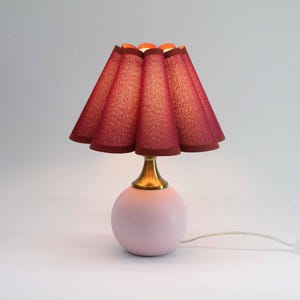 Peut inclure: Une petite lampe de table rose avec une base ronde et une tige dorée. L'abat-jour est rouge foncé avec un motif texturé et un bord festonné. La lampe a un cordon blanc.