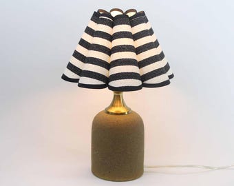 Puede incluir: Lámpara de mesa decorativa con base beige texturizada y cuello de latón. La pantalla es plisada, con rayas horizontales negras y blancas alternas. La lámpara está sobre un fondo blanco.
