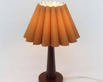 May include: A small, brown table lamp with a pleated lampshade. The lampshade is a light brown color and has a flower-like shape. The base is dark brown and round. The lamp is on a white background.