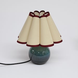 May include: A table lamp with a pleated cream-colored lampshade with a burgundy trim. The lamp base is a dark blue and green ceramic sphere.