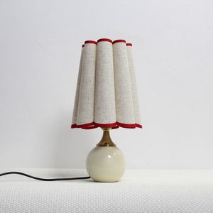 May include: A white table lamp with a pleated linen shade trimmed in red. The lamp has a round base and a gold-colored metal stem.