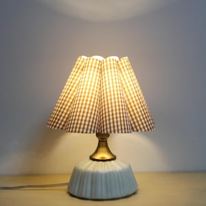 Può includere: Una lampada da tavolo in ceramica bianca con un paralume a quadri marroni e bianchi. Il paralume ha un design plissettato ed è acceso.