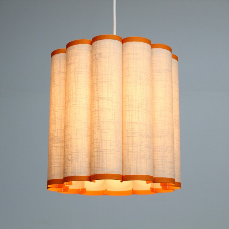 May include: A cylindrical pendant light fixture with a white linen shade and orange trim. The shade is composed of vertical, tube-like segments, creating a textured appearance. The light fixture hangs from a white cord.