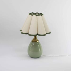 May include: A decorative table lamp with a sage green ceramic base and a brass neck. The lampshade is cream-colored with a green trim. The lamp has a unique, pleated design and a white cord.