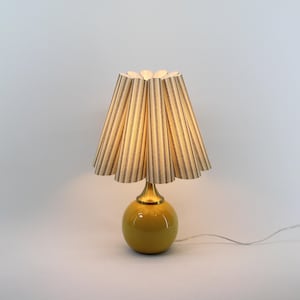 May include: A yellow ceramic table lamp with a striped lampshade. The lamp has a round base and a brass-coloured neck. The lampshade is beige with vertical brown stripes. The lamp is plugged in and lit, casting a warm glow.