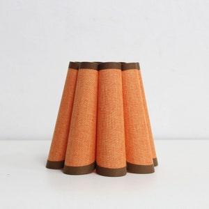 Peut inclure: Abat-jour en tissu orange avec une bordure marron. L'abat-jour a un design plissé, chaque pli formant une forme de cône. L'abat-jour est posé sur une surface blanche.