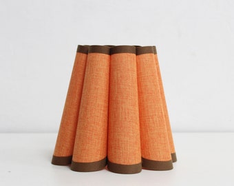 May include: Orange fabric lampshade with a brown trim. The lampshade has a pleated design, with each pleat forming a cone shape. The lampshade is sitting on a white surface.