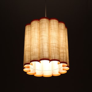 May include: A cylindrical pendant light fixture with a series of vertical, textured, beige fabric columns. The top and bottom edges are trimmed with a dark red material. The interior glows with warm light, creating a soft ambiance. The fixture hangs from a white cord.