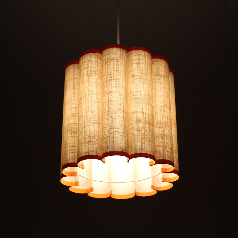 May include: A cylindrical pendant light fixture with a series of vertical, textured, beige fabric columns. The top and bottom edges are trimmed with a dark red material. The interior glows with warm light, creating a soft ambiance. The fixture hangs from a white cord.