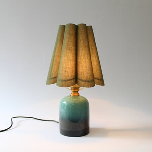May include: A green pleated lampshade sits atop a teal and black ceramic lamp base. The lamp has a gold metal base and a black cord.