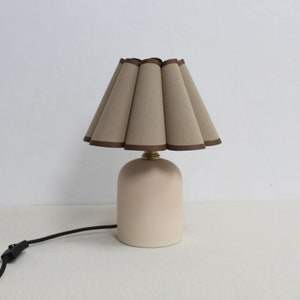 May include: A beige ceramic table lamp with a pleated beige fabric lampshade with a brown trim.