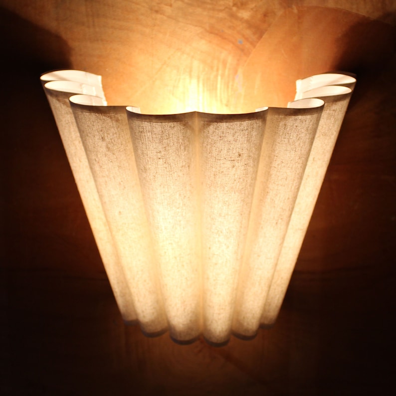 May include: A decorative wall sconce with a unique, scalloped design. The sconce is made of a light-colored, textured fabric, and the light shines through the fabric, creating a warm glow. The sconce is mounted on a dark brown wall.