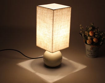 Può includere: Una lampada da tavolo bianca con un paralume quadrato in lino e una base rotonda bianca. La lampada è accesa e proietta una luce calda.
