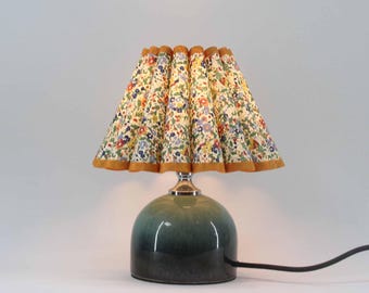 May include: A small table lamp with a floral patterned lampshade. The lampshade is cream-colored with a pattern of colorful flowers and a gold trim. The base is a rounded, two-toned green ceramic.