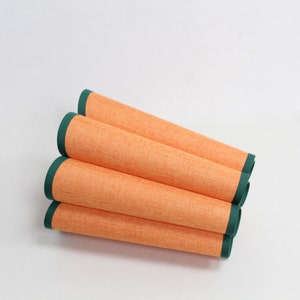 May include: Four orange placemats with a dark green border are rolled up together. The placemats have a textured surface.