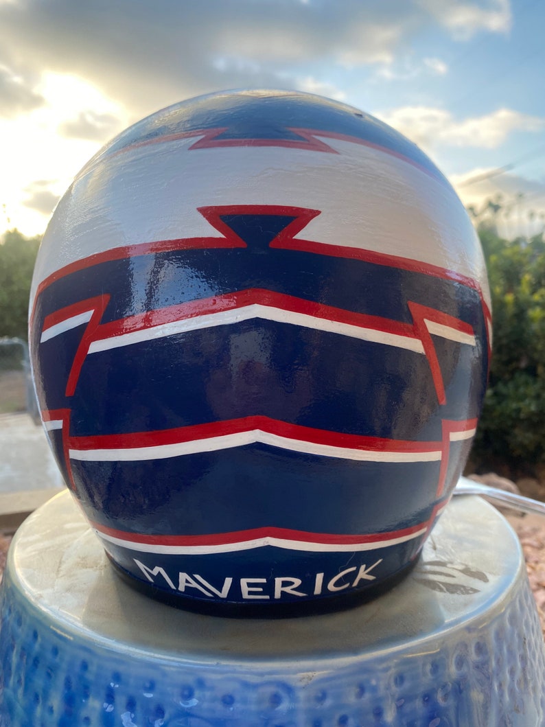 May include: A blue, white, and red painted motorcycle helmet with the word "MAVERICK" written in white on the bottom.