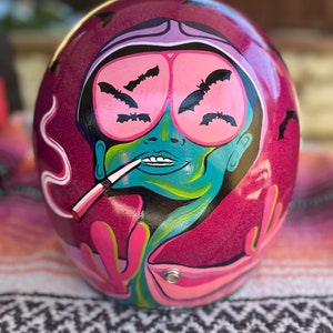 May include: A pink motorcycle helmet with a painted design of a person with pink goggles and bats. The person is smoking a cigarette and has a green face.