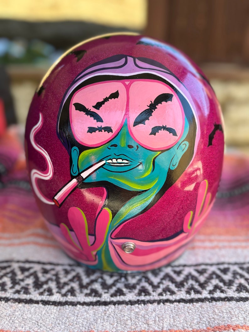 May include: A pink motorcycle helmet with a painted design of a person with pink goggles and bats. The person is smoking a cigarette and has a green face.