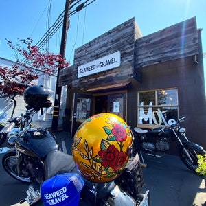 May include: A black motorcycle with a blue and white baseball cap that says "Seaweed & Gravel" on it. A gold helmet with a floral design is on the motorcycle. The motorcycle is parked in front of a store with a sign that says "Seaweed & Gravel".