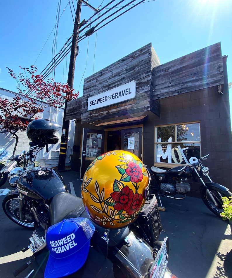 May include: A black motorcycle with a blue and white baseball cap that says "Seaweed & Gravel" on it. A gold helmet with a floral design is on the motorcycle. The motorcycle is parked in front of a store with a sign that says "Seaweed & Gravel".