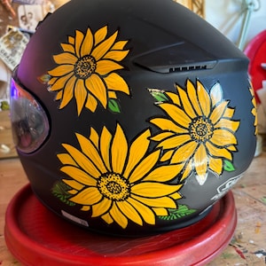 May include: A black motorcycle helmet with hand-painted yellow sunflowers. The helmet is sitting on a red circular stand.