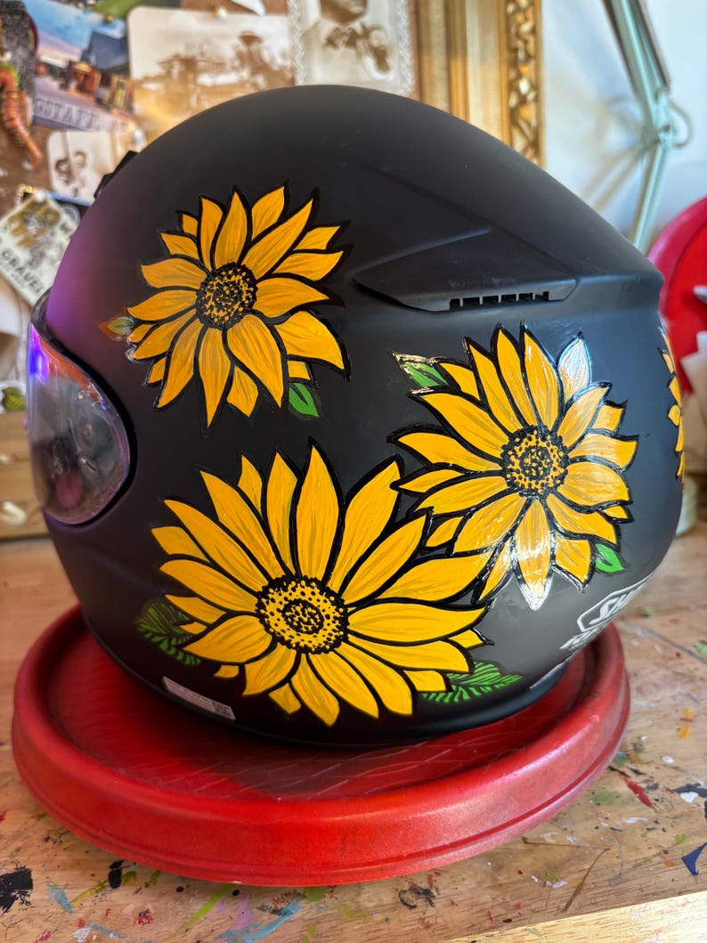 May include: A black motorcycle helmet with hand-painted yellow sunflowers. The helmet is sitting on a red circular stand.