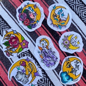 May include: Seven colorful crescent moon illustrations with various designs, including flowers, candles, and a face. The illustrations are painted on white paper and have a whimsical, vintage style.