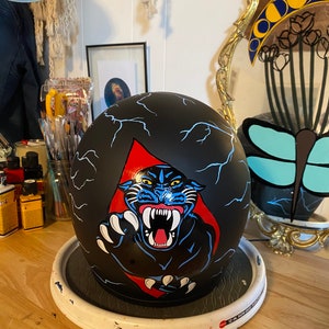 May include: Black motorcycle helmet with a hand-painted design of a black panther with blue and red accents. The panther is depicted with a fierce expression and sharp teeth. The helmet is on a white circular stand.