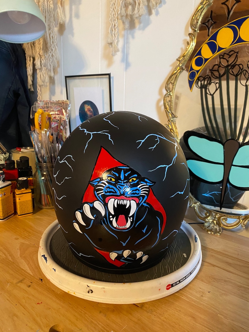 May include: Black motorcycle helmet with a hand-painted design of a black panther with blue and red accents. The panther is depicted with a fierce expression and sharp teeth. The helmet is on a white circular stand.