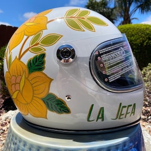 May include: White motorcycle helmet with a floral design in yellow, green, and pink. The helmet has a clear visor and the words "LA JEFA" are written in green on the bottom.