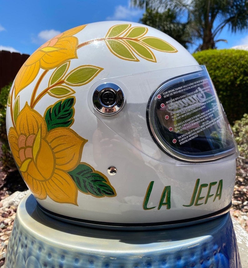 May include: White motorcycle helmet with a floral design in yellow, green, and pink. The helmet has a clear visor and the words "LA JEFA" are written in green on the bottom.