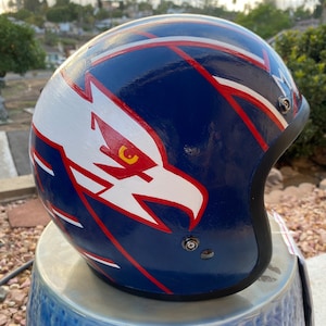 May include: A blue, white, and red motorcycle helmet with an eagle design. The eagle has a yellow eye and is facing to the right. The helmet is sitting on a blue and white ceramic stool.