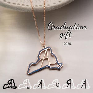 May include: Rose gold necklace with a pendant shaped like a state outline, featuring the letter "A" in the center. The pendant has a dark blue border. The words "Graduation gift 2026" are written in cursive. Several outline designs of the pendant are at the bottom.