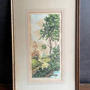 May include: Framed landscape art print featuring a serene countryside scene. The artwork depicts a path leading to a small house, with trees and a river. The print is in a gold-toned frame, adding a classic touch to the artwork.