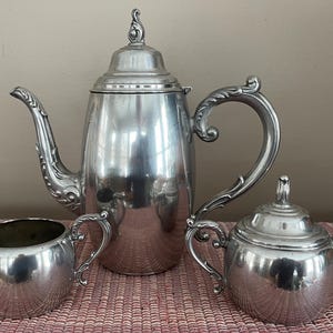 May include: A silver tea set with a teapot, creamer, and sugar bowl. The teapot has a curved spout and ornate handle. The creamer and sugar bowl have matching handles. The set is displayed on a pink surface.