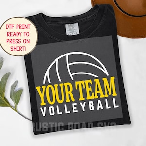 Könnte beinhalten: Schwarzes T-Shirt mit einem Volleyball-Grafik. Das Design zeigt einen weißen Volleyball und die Worte "YOUR TEAM VOLLEYBALL" in Gelb und Weiß. Das Shirt hat auch einen runden Aufkleber mit der Aufschrift "DTF PRINT READY TO PRESS ON SHIRT!"