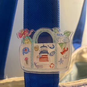 May include: A clear acrylic charm featuring a watercolor illustration of a tote bag with nautical-themed elements. The charm includes a lighthouse, lobster, ice cream cone, and a whale, attached to a blue tote bag strap.