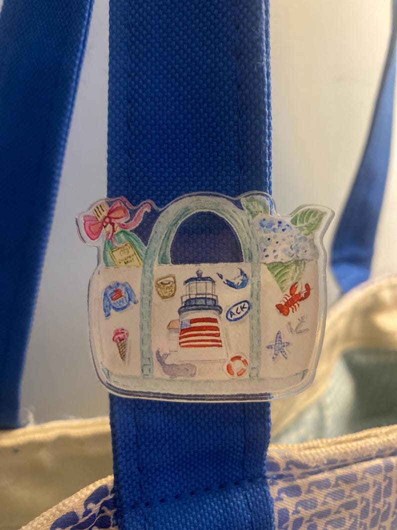 May include: A clear acrylic charm featuring a watercolor illustration of a tote bag with nautical-themed elements. The charm includes a lighthouse, lobster, ice cream cone, and a whale, attached to a blue tote bag strap.